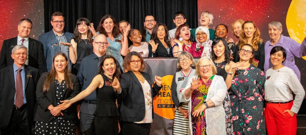 WayAhead staff, board and volunteers at the 2019 Mental Health Matters awards. Everyone waving, laughing or smiling while standing around the lectern on the stage.